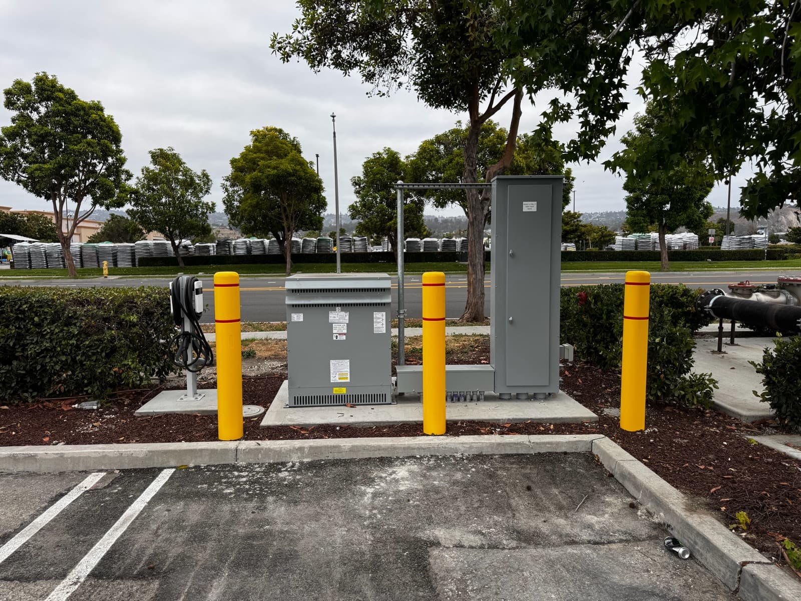 Transformer and panel enclosure with a roadside charging pedestal behind yellow bollards